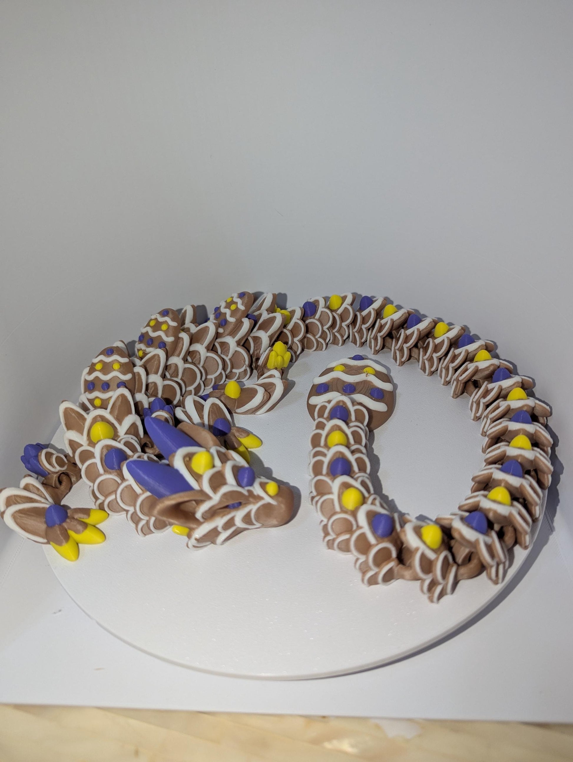 Articulated Easter Egg Dragon. Featuring four colors (Brown,white,yellow,and purple). Curled up with a large Easter egg tail. side profile